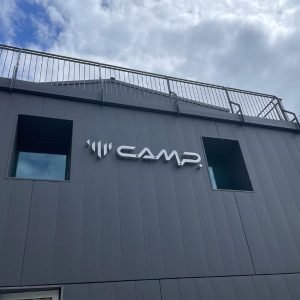 Blick auf den weißen Schriftzug C.A.M.P. und das Piktogramm am grauen Firmengebäude, darüber ein Geländer und wolkiger Himmerl