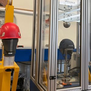 Grauer Kletterhelm in der Aufpralltestung, daneben roter Industrieschutzhelm